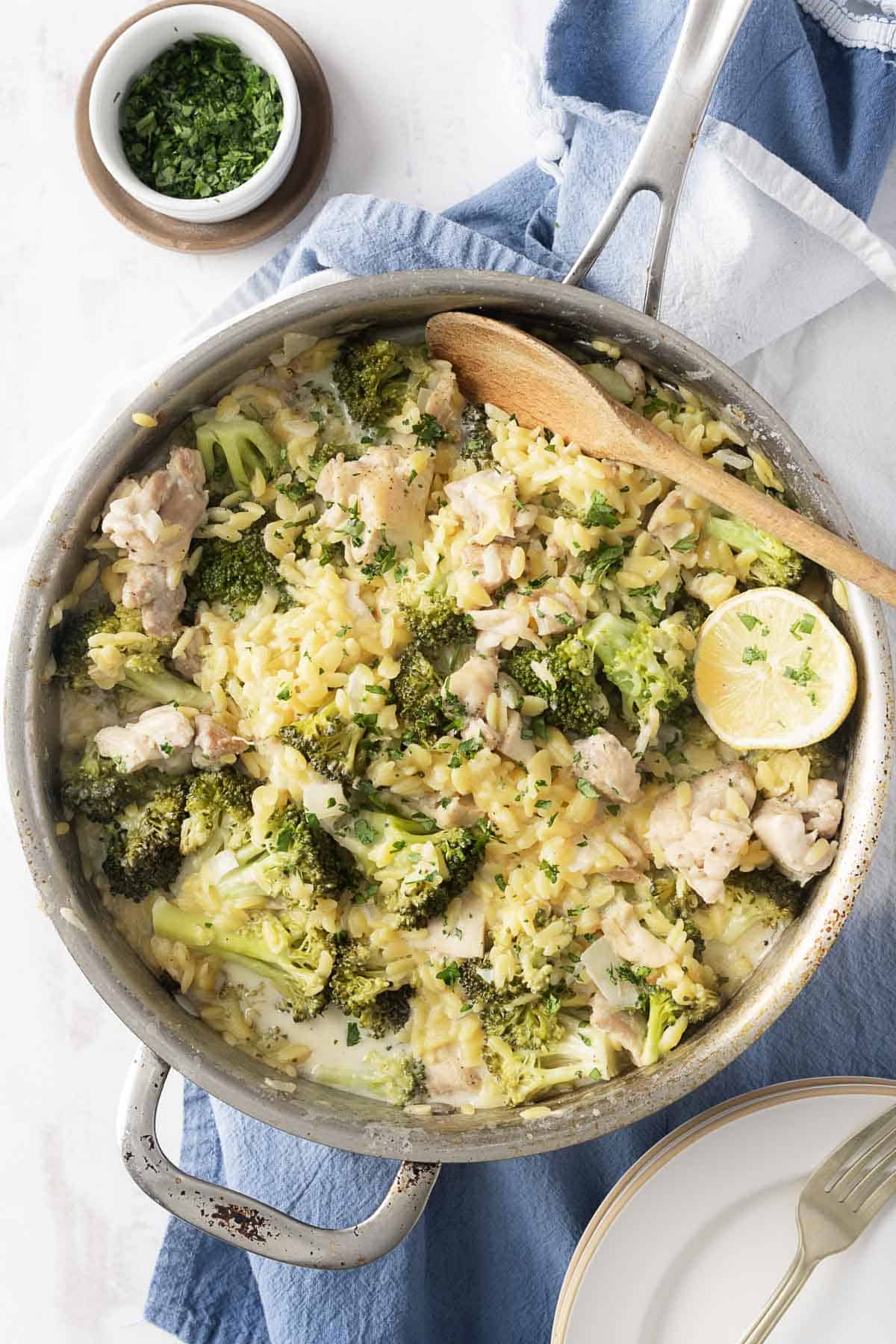 A one-pot dish of creamy pasta with broccoli, tender chicken, and a hint of Boursin cheese, garnished with lemon and herbs, rests on a blue towel.