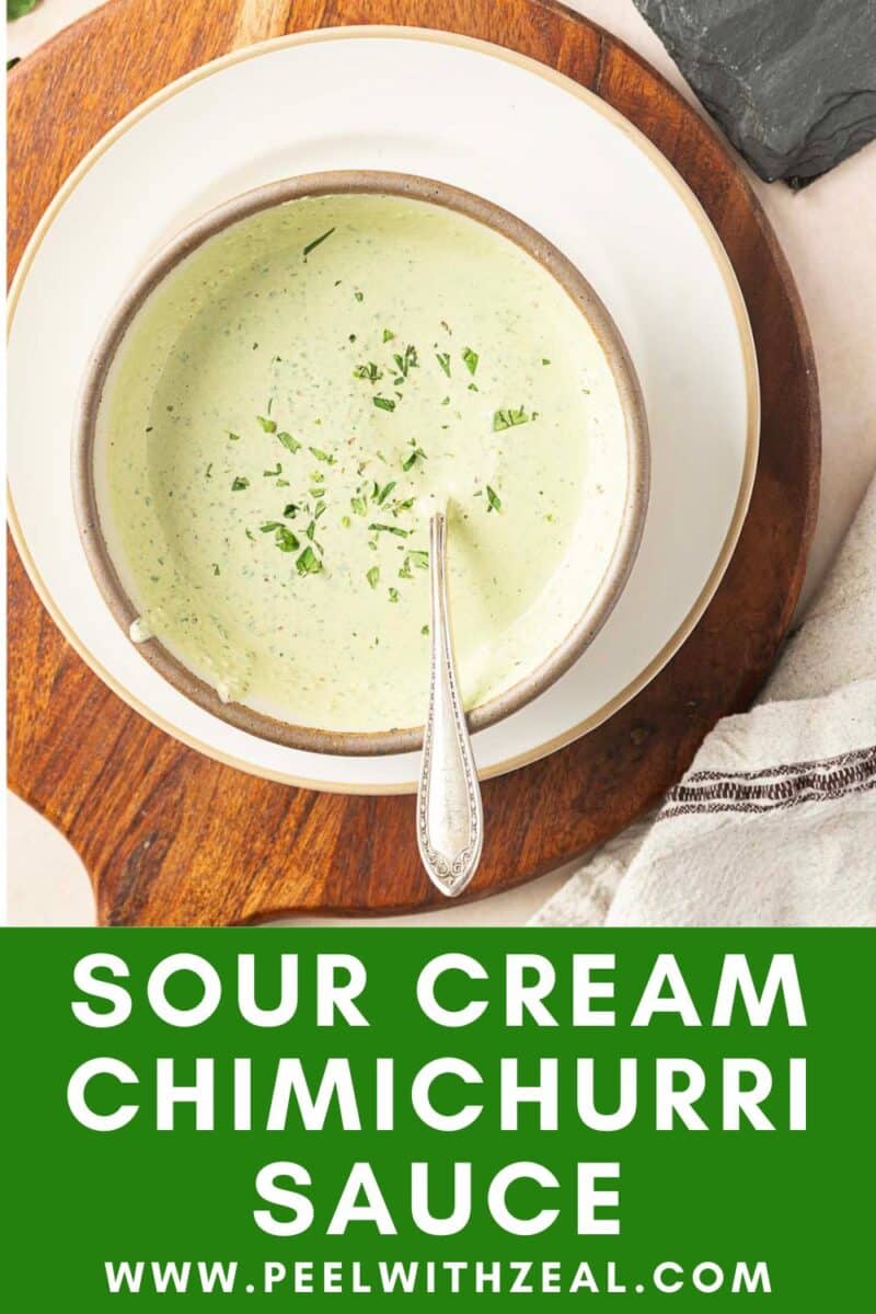 A bowl of creamy chimichurri sauce with a spoon, garnished with herbs, sits elegantly on a wooden board.