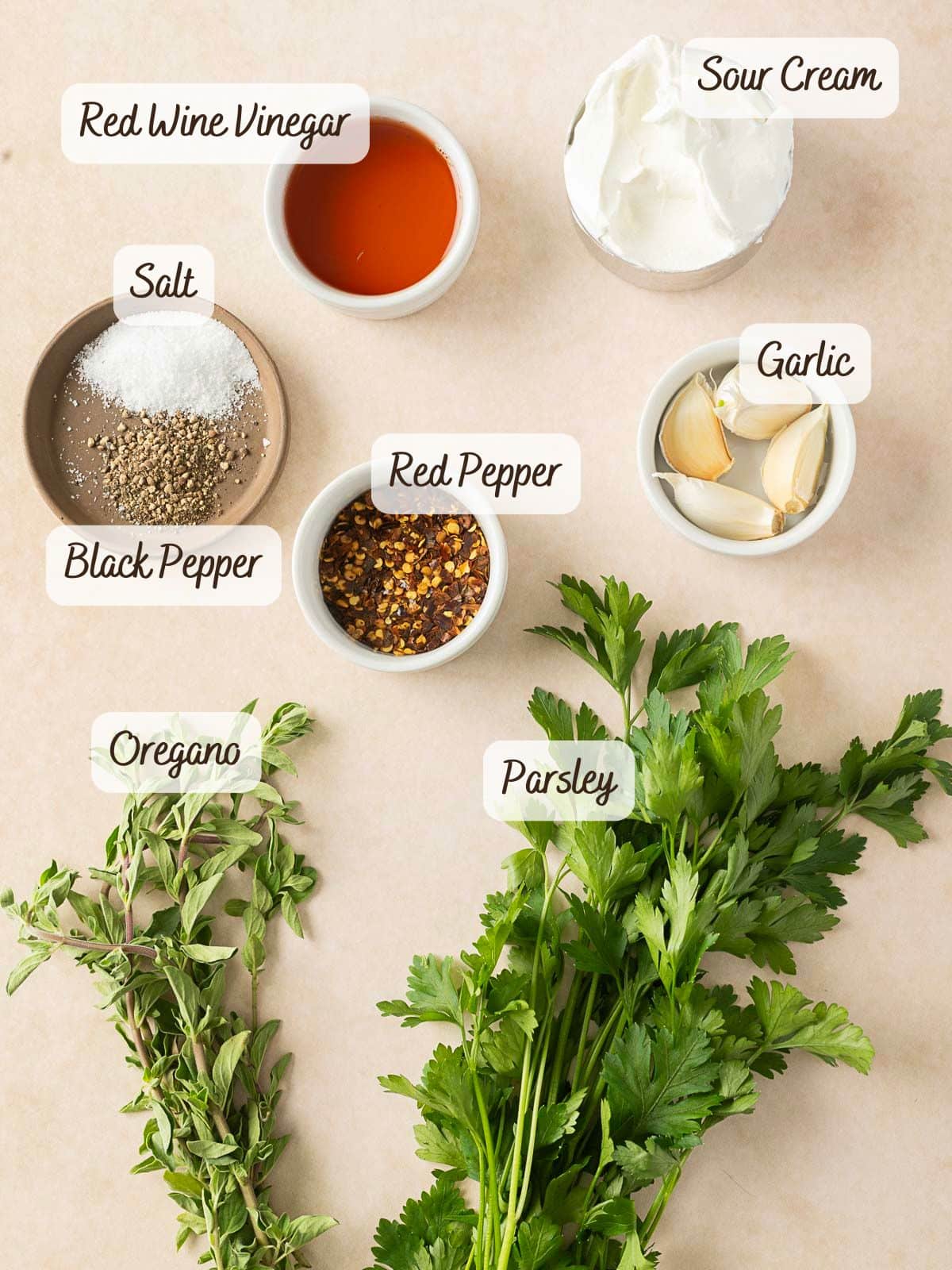 A variety of ingredients labeled on a surface: red wine vinegar, sour cream, salt, black pepper, red pepper flakes, garlic cloves, oregano and parsley – the perfect mix for a creamy chimichurri sauce.