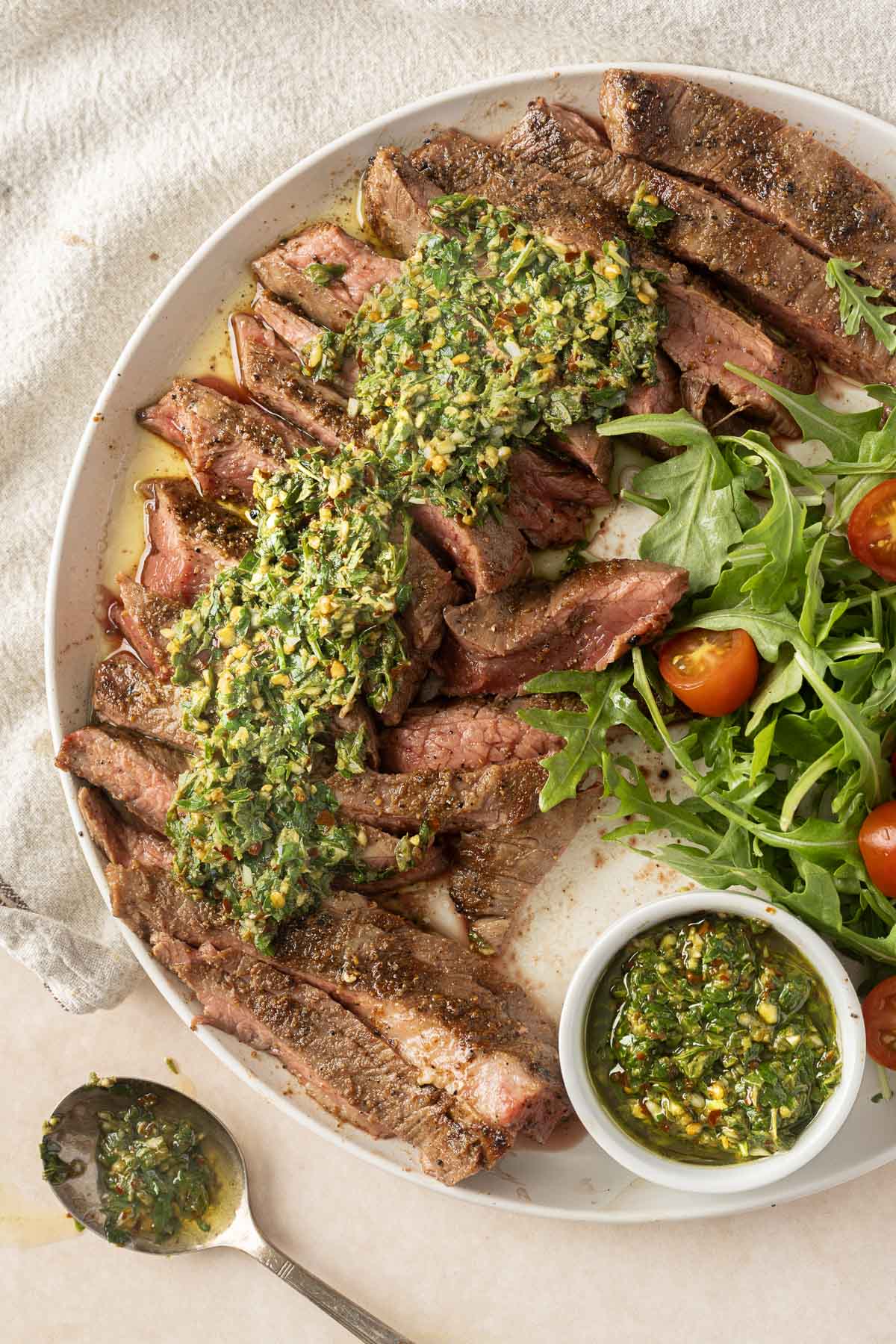 a delectable grilled flank steak topped with vibrant green chimichurri sauce, perfectly paired with fresh arugula and cherry tomatoes for a mouthwatering dish.