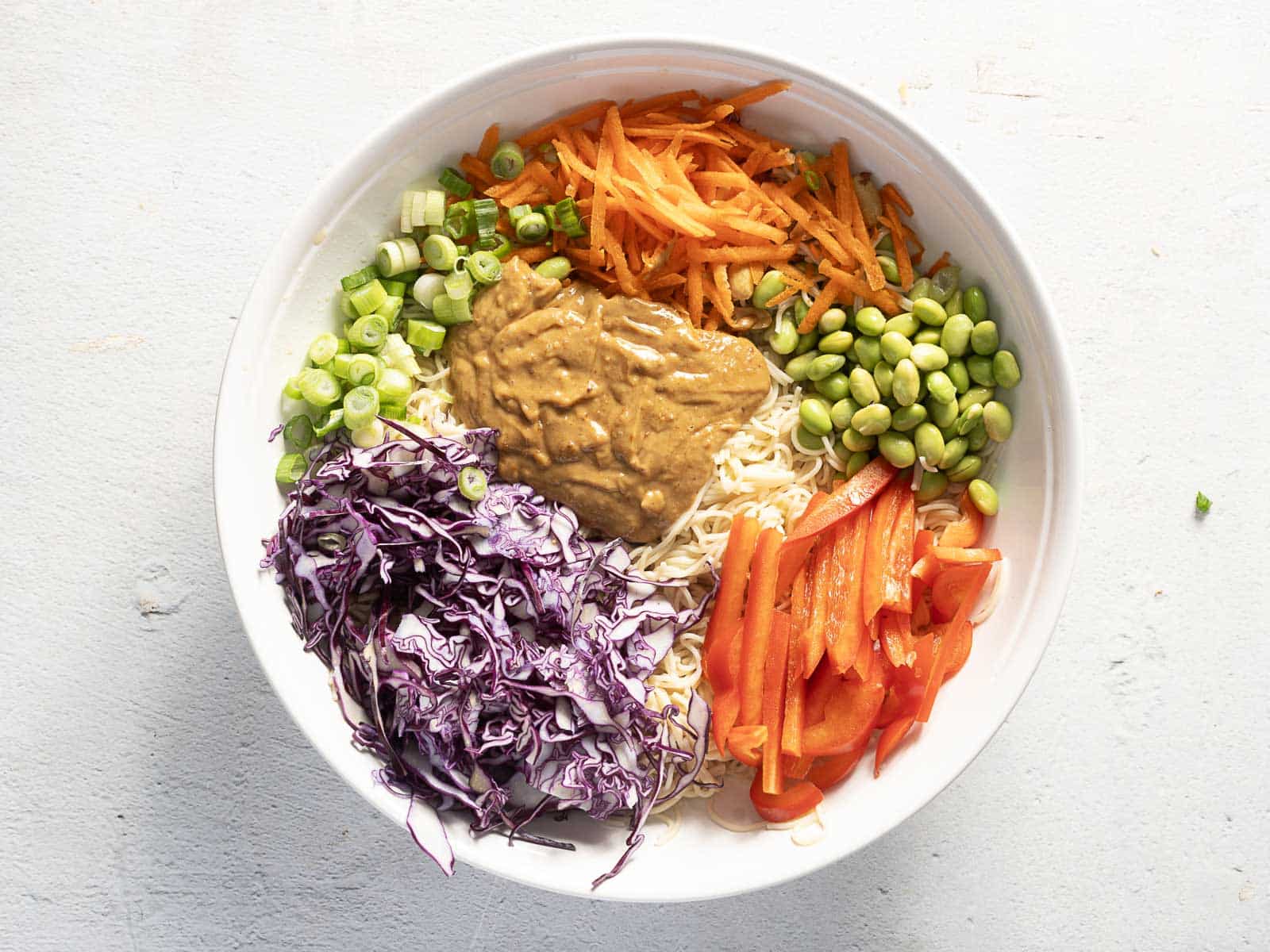 A white bowl with cold ramen noodles, shredded carrots, edamame, red bell peppers, sliced green onions, and purple cabbage is topped with a delectable brown sauce that's reminiscent of peanut dressing.