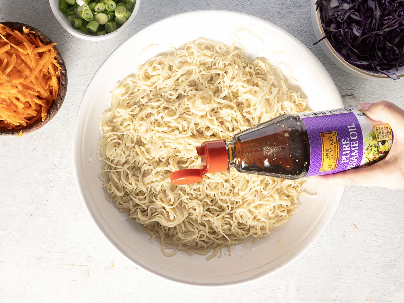 A person drizzles sesame oil over a bowl of cold ramen, transforming it into a refreshing noodle salad. Shredded carrots, green onions, and cabbage await in separate bowls nearby.