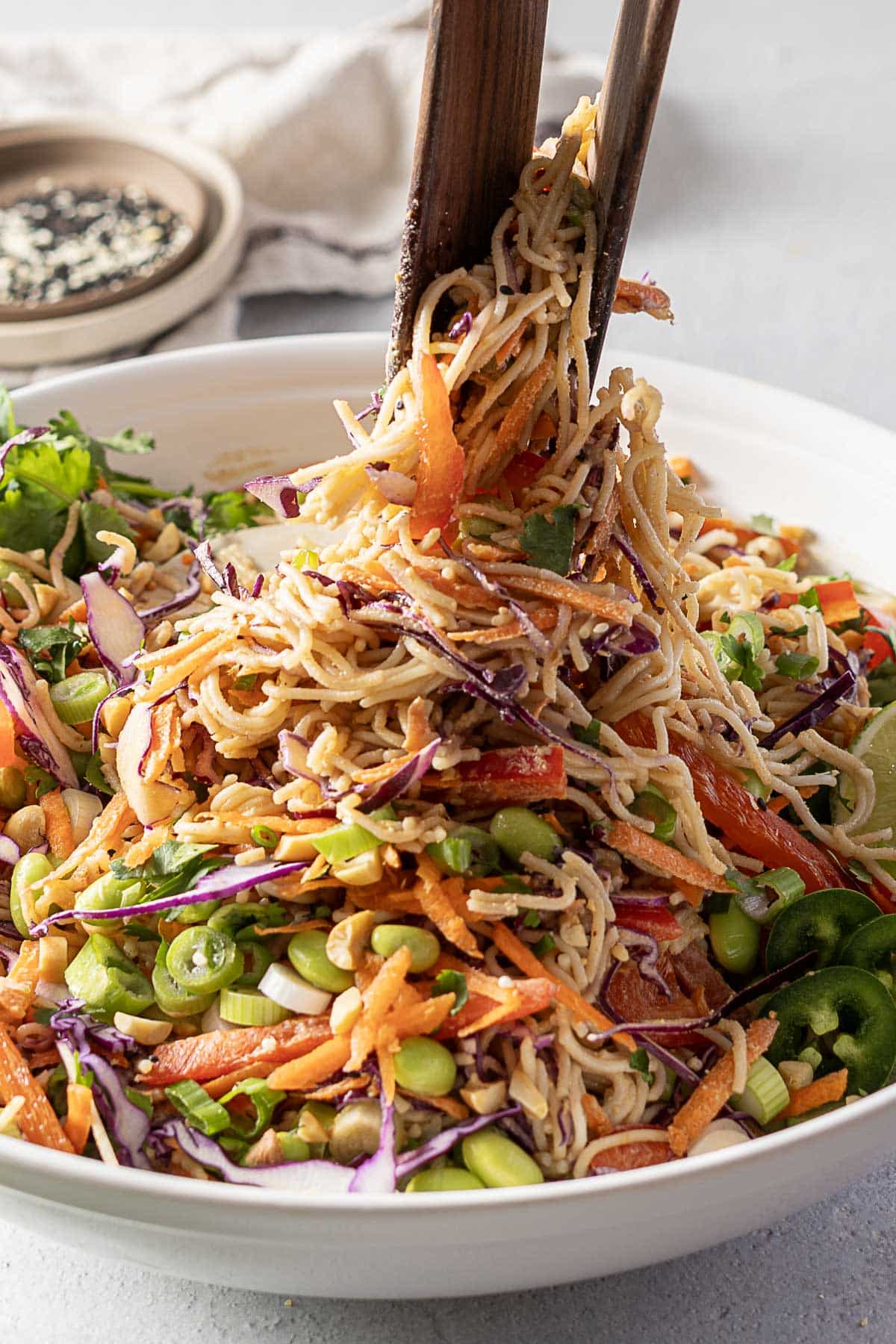 tongs lift a portion of noodle salad dressed in a rich peanut dressing from a white bowl, bursting with vibrant vegetables and greens.