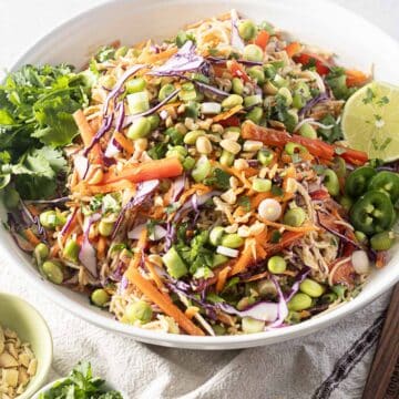 A bowl of cold ramen noodle salad with shredded cabbage, carrots, edamame, sliced jalapeños, chopped peanuts, and fresh cilantro. It's drizzled with a delicious peanut dressing and garnished with lime and green onions.