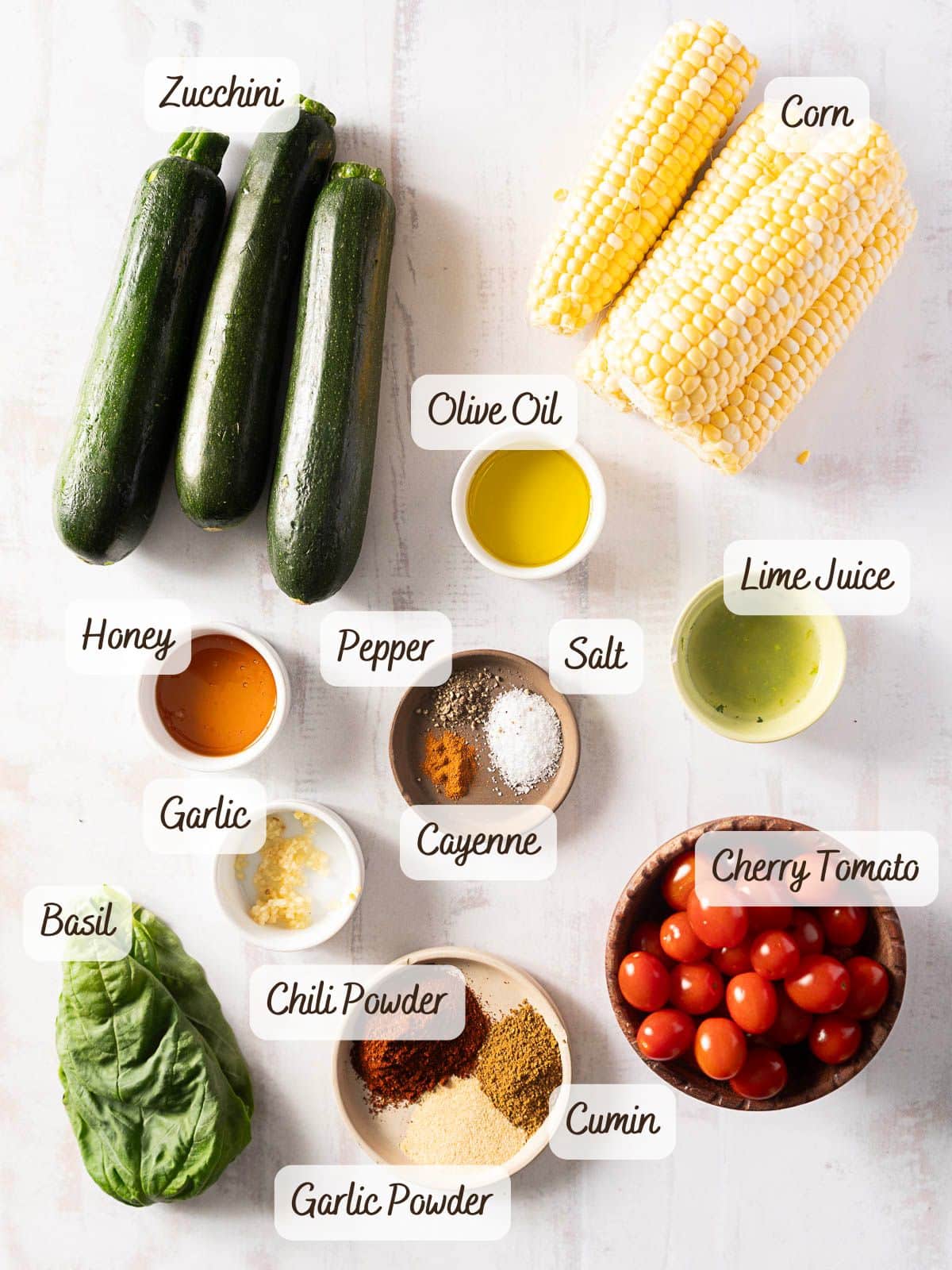 Ingredients for a refreshing corn and zucchini salad on a white surface: zucchini, corn, olive oil, lime juice, cherry tomatoes, basil, honey, garlic, pepper, salt, cayenne, chili powder, garlic powder, and cumin.