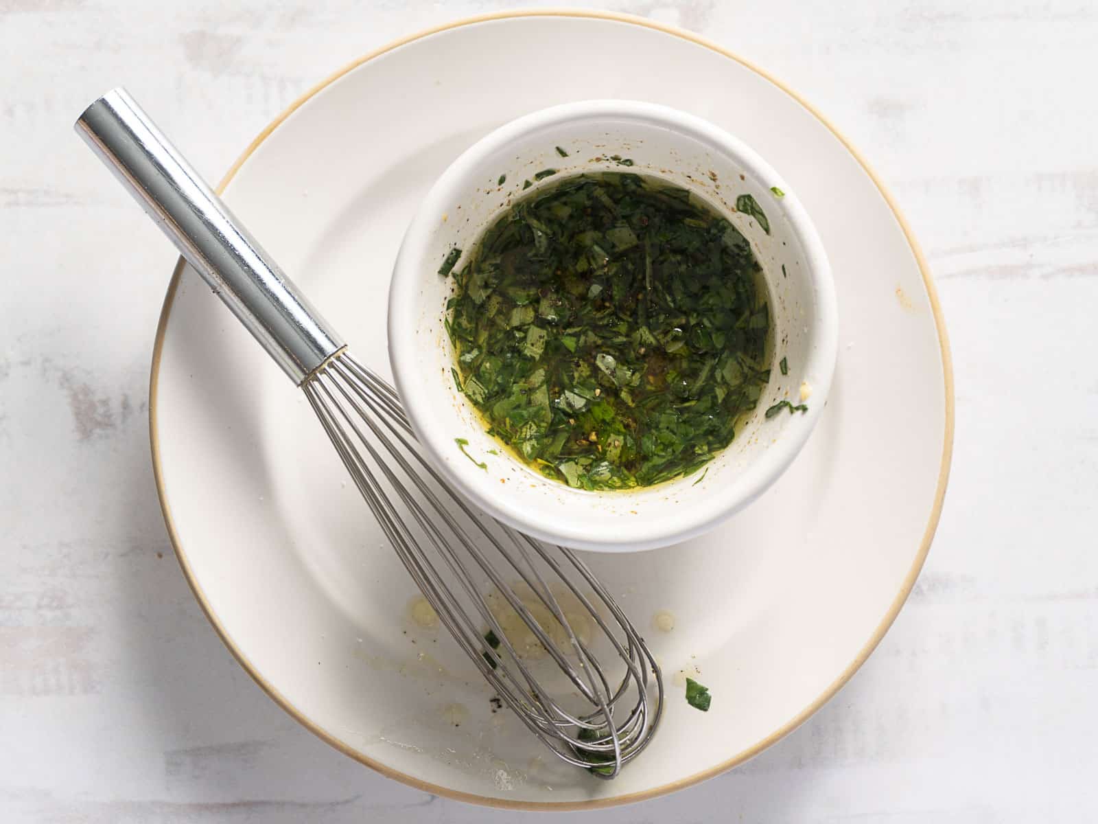 A metal whisk rests on a white plate beside a bowl of chopped herbs mixed in oil, complementing the fresh corn and zucchini salad prepared to perfection.