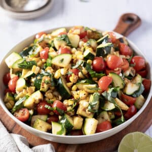 A vibrant corn and zucchini salad sits invitingly on a wooden board, boasting cherry tomatoes and fresh basil. A lime wedge awaits in the foreground, hinting at a zesty finish, while a touch of salt lingers in the background.