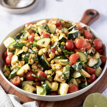 A vibrant corn and zucchini salad sits invitingly on a wooden board, boasting cherry tomatoes and fresh basil. A lime wedge awaits in the foreground, hinting at a zesty finish, while a touch of salt lingers in the background.