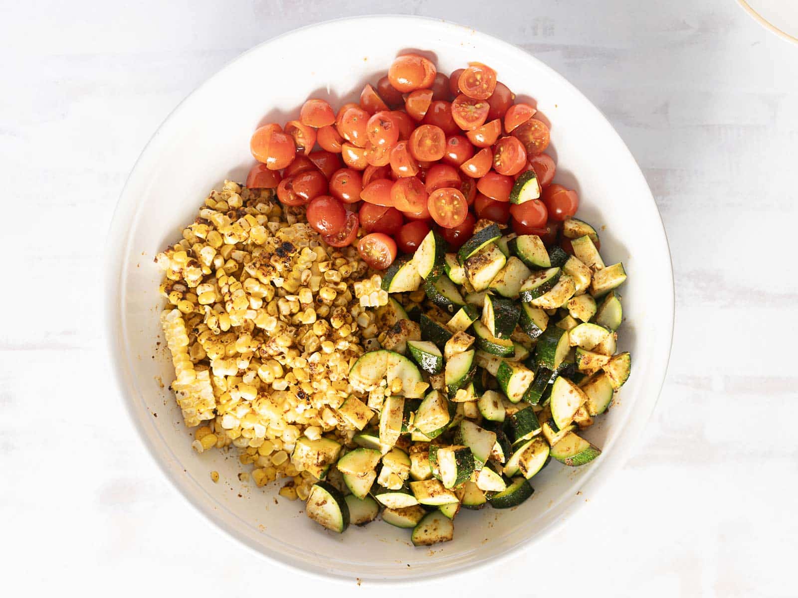 A vibrant corn and zucchini salad gleams with grilled corn, sweet cherry tomatoes, and diced zucchini in a bowl against a crisp white background.