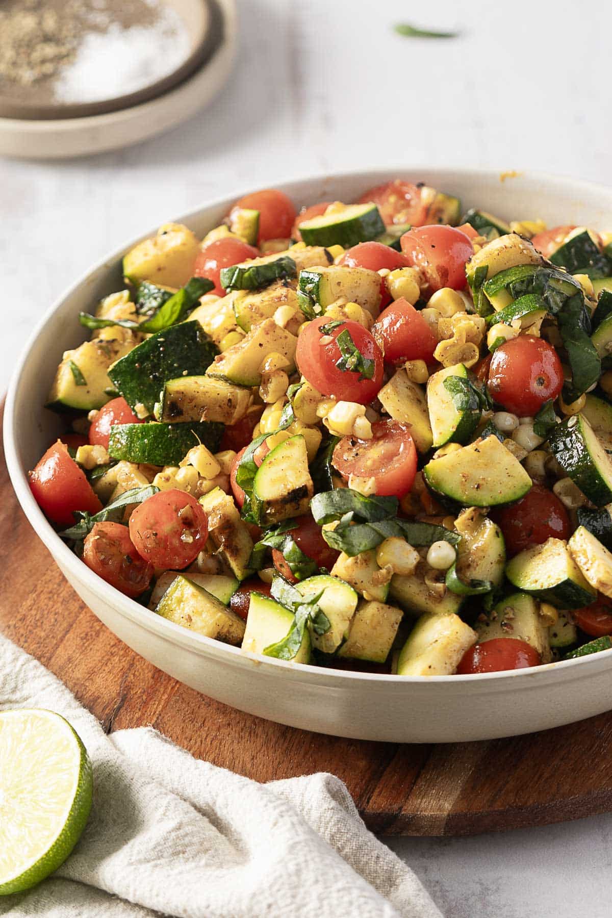 A corn and zucchini salad, featuring cherry tomatoes and basil, is artfully arranged in a bowl on a wooden board. Nearby, a fresh lime slice and white cloth add a touch of rustic simplicity.