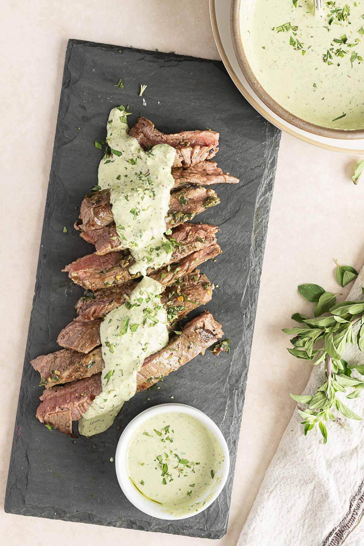Sliced steak on a slate board topped with creamy chimichurri sauce, garnished with herbs. A small bowl of this aromatic sauce is next to the steak. A napkin and fresh herbs are on the side.