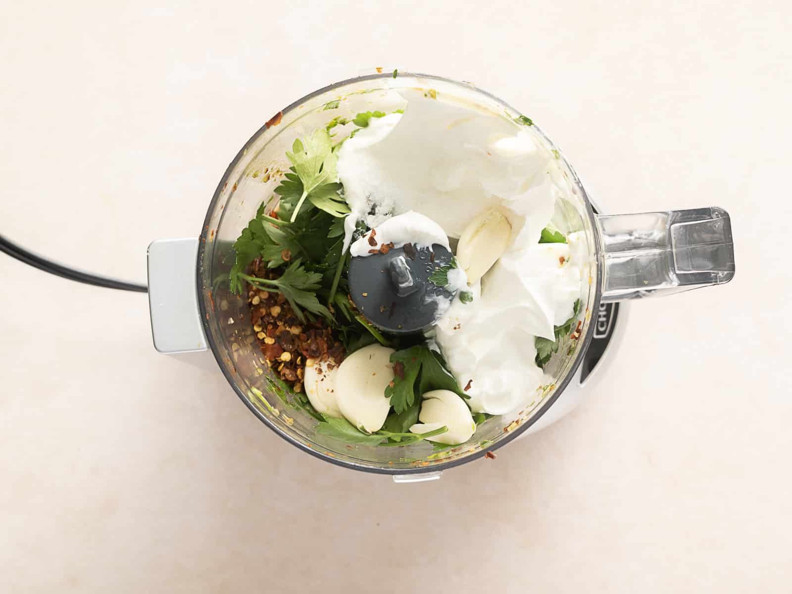 A creamy blend of cilantro, garlic cloves, yogurt, and red pepper flakes sits invitingly in the food processor, reminiscent of a modern twist on Chimichurri Sauce.