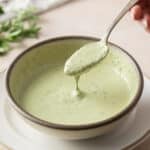 A hand holds a spoon above a bowl of creamy chimichurri sauce, garnished with fresh herbs, on a light-colored surface.