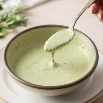 A hand holds a spoon above a bowl of creamy chimichurri sauce, garnished with fresh herbs, on a light-colored surface.