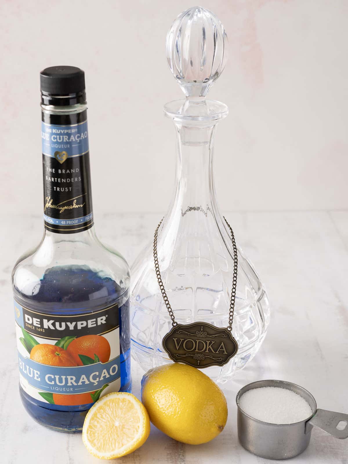 a Blue Curaçao bottle, a vodka decanter, a zesty lemon, half sliced for garnish, and sugar perfectly measured in a cup.