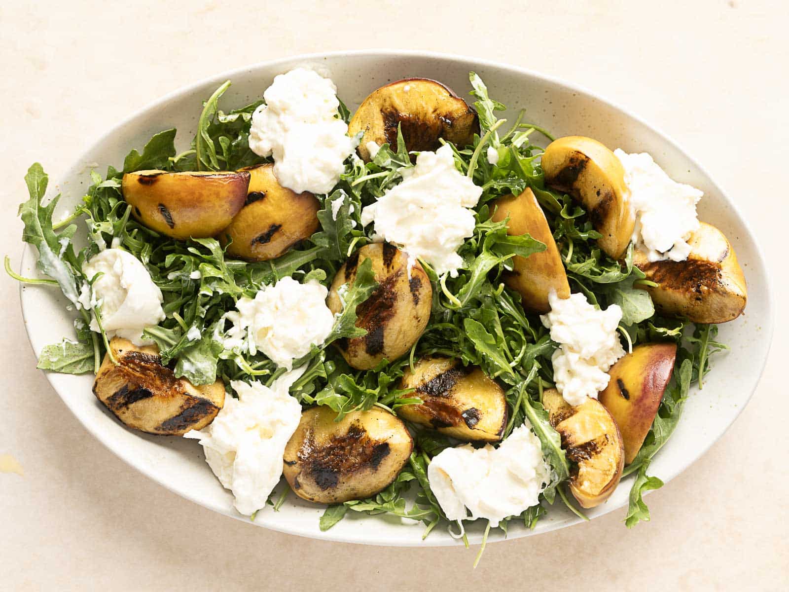 Oval plate showcasing a delightful grilled peach salad, featuring succulent peach halves paired with creamy burrata cheese and fresh arugula on a light surface.