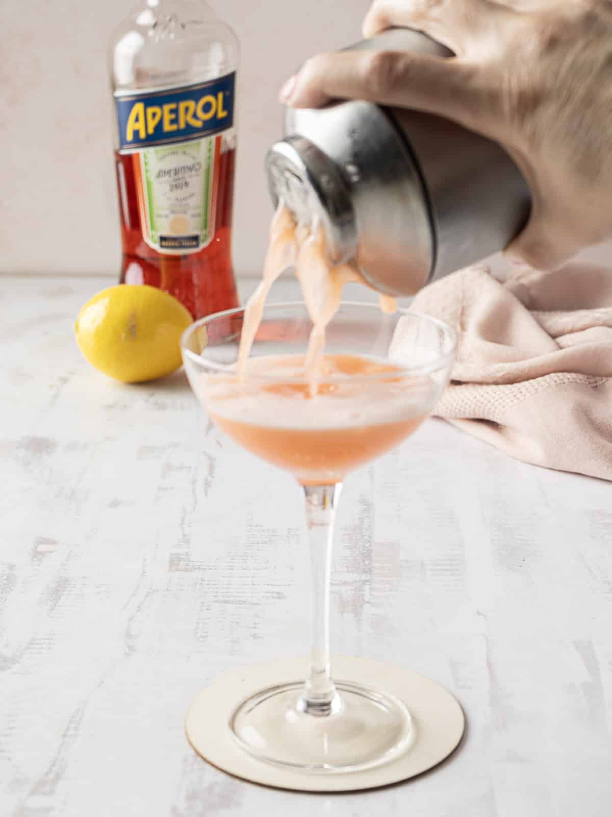 A hand pours an orange cocktail from a shaker into a glass. A bottle of Aperol and a lemon are in the background.