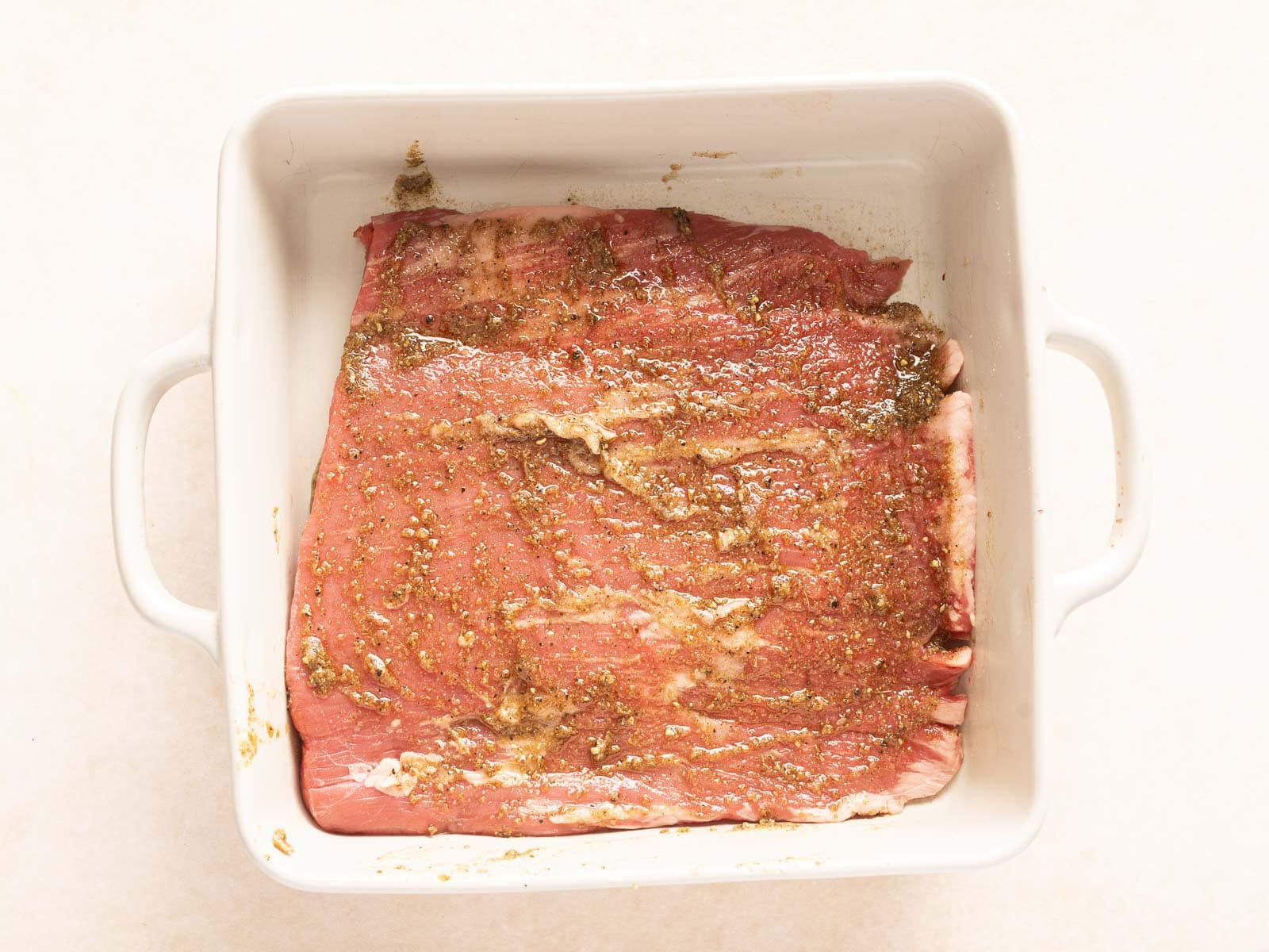 Raw marinated meat, destined to become a succulent grilled flank steak, rests enticingly in a white baking dish on a light surface.