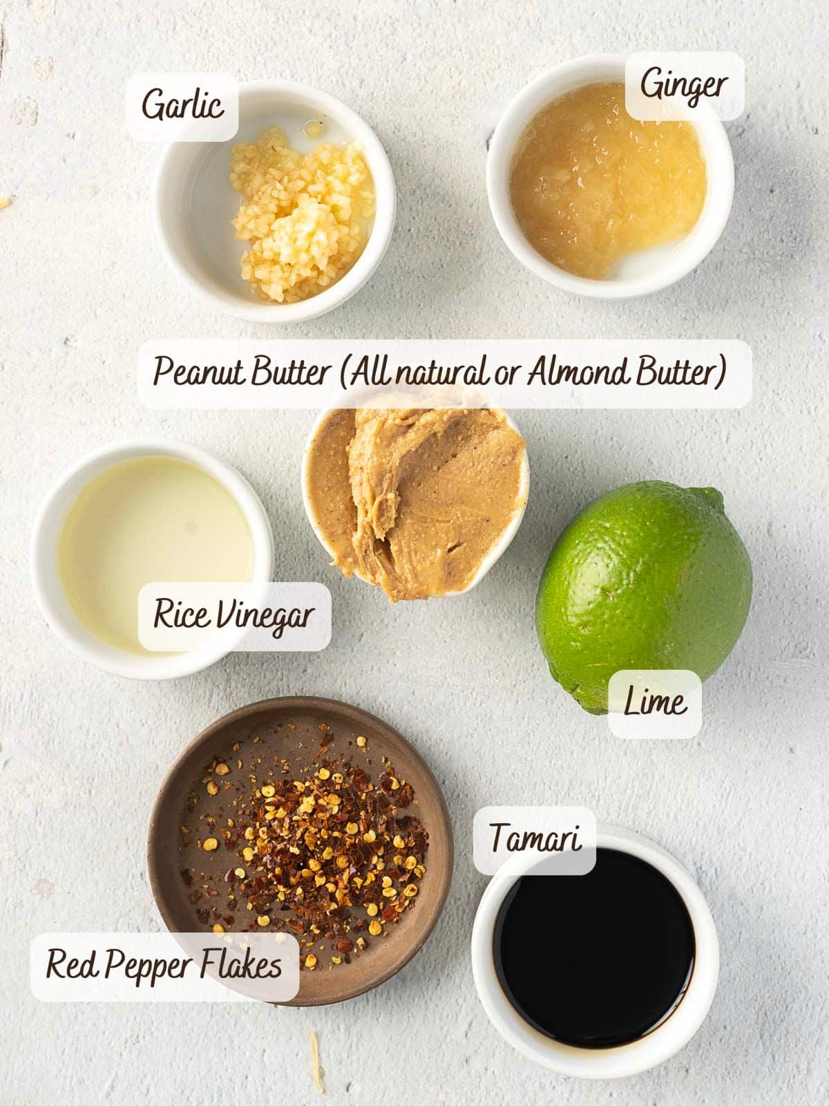 Ingredients in small bowls arranged on a surface: minced garlic, grated ginger, natural peanut butter, rice vinegar, whole lime, red pepper flakes, and tamari.