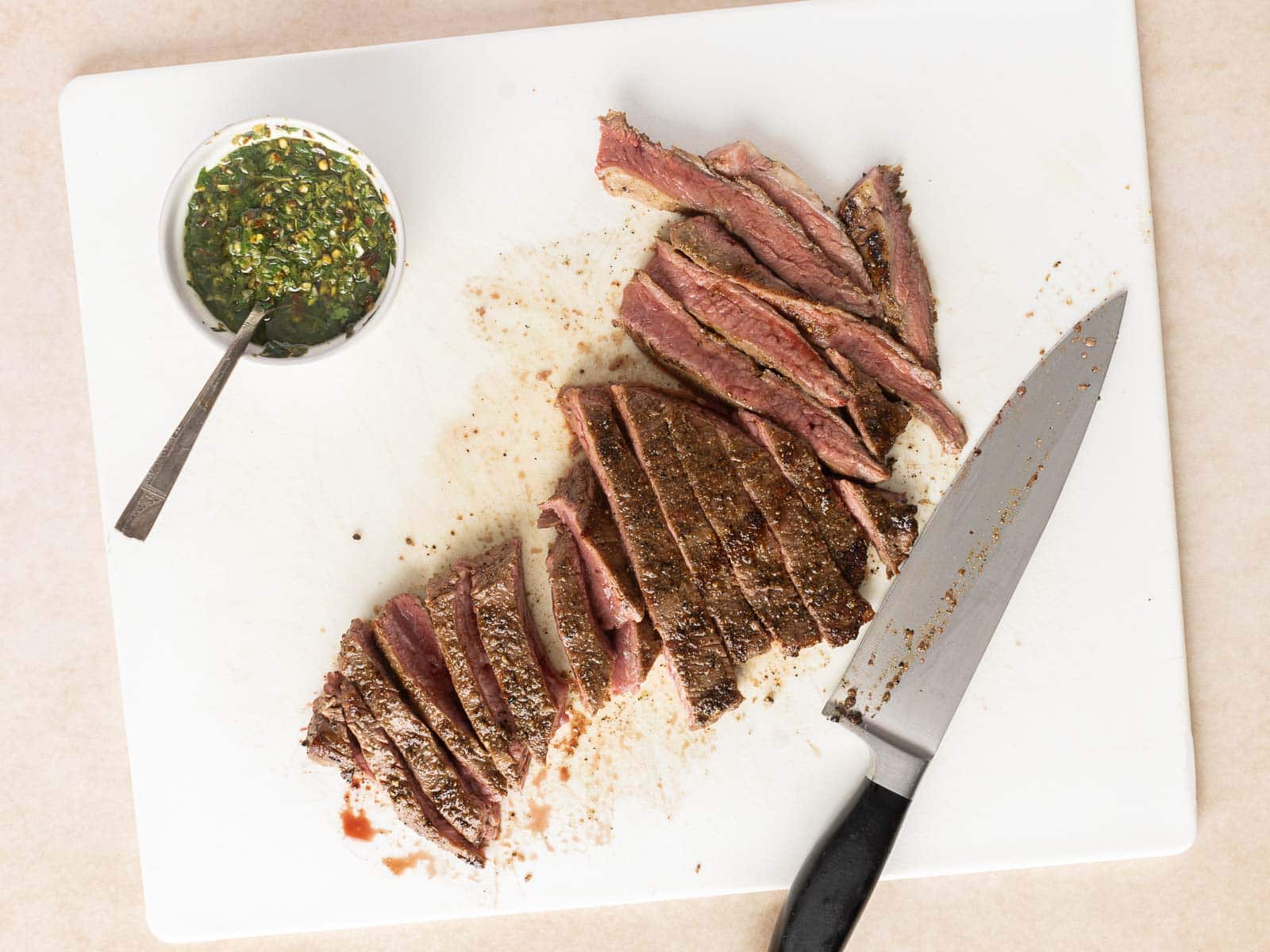 Grilled flank steak is artfully sliced on a cutting board, accompanied by a sharp knife and a vibrant bowl of chimichurri sauce.