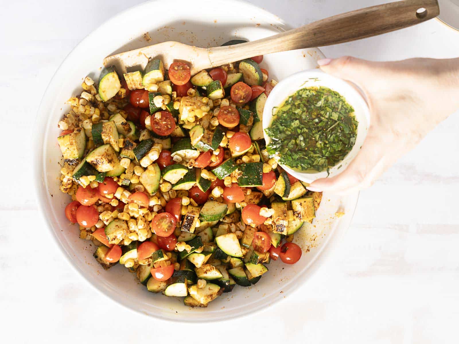 A bowl of corn and zucchini salad features grilled corn, zucchini, and cherry tomatoes. A hand hovers above with a small bowl of green dressing, ready to drizzle.