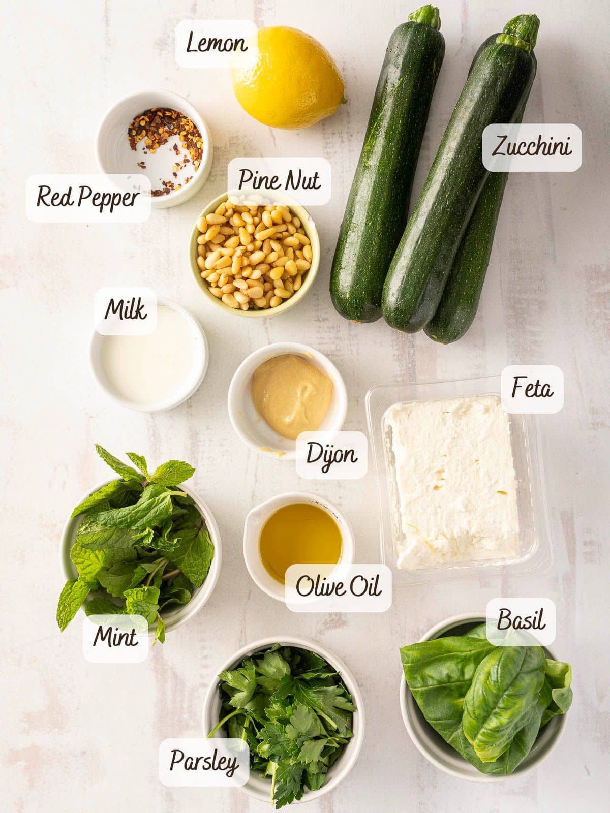 On a pristine white surface, you'll find the makings of a refreshing zucchini salad: fresh zucchini, crumbled feta, zesty lemon, and an assortment of aromatic herbs and seasonings.