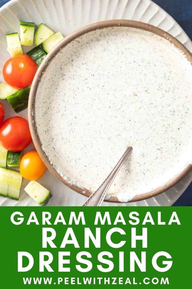 A bowl of Indian salad dressing, garam masala ranch style, with a spoon, surrounded by cherry tomatoes and cucumber pieces.