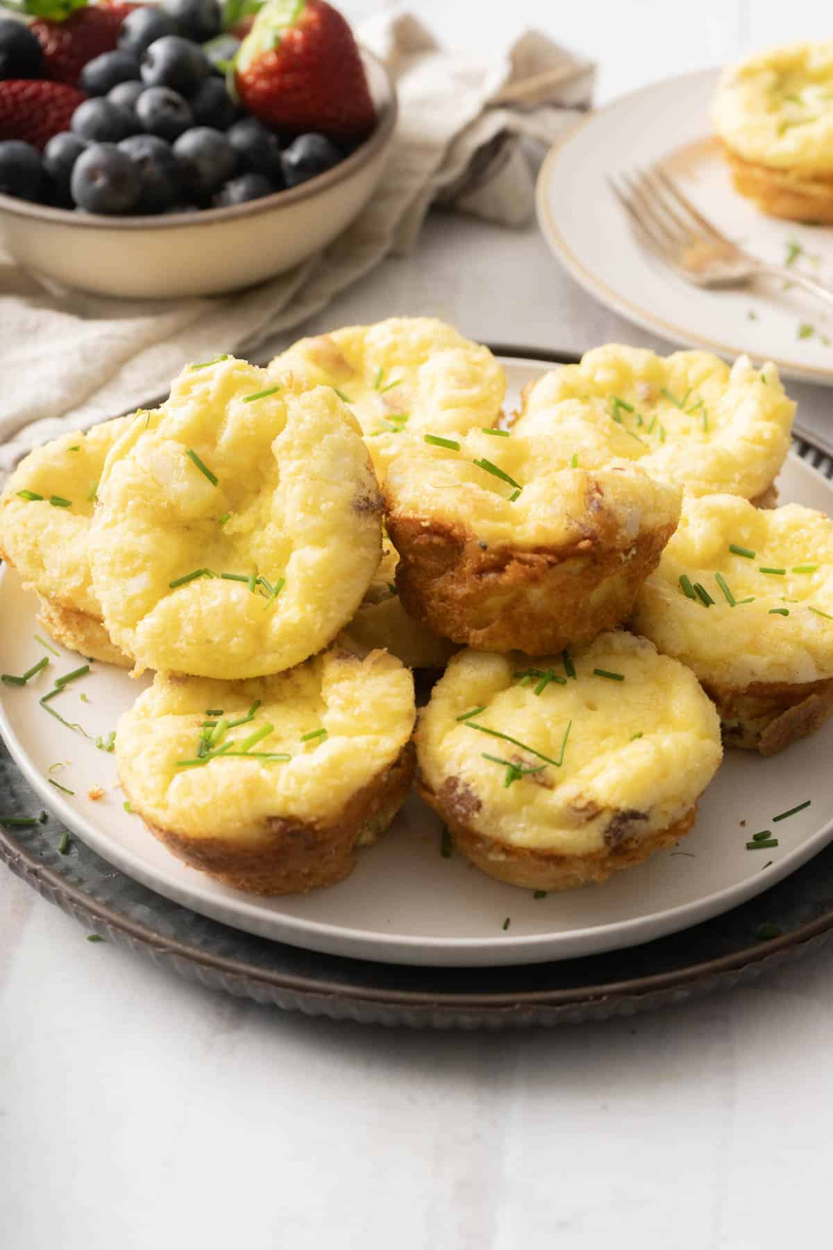 A plate of quiche muffins garnished with chives sits elegantly beside a bowl of mixed berries on the table.