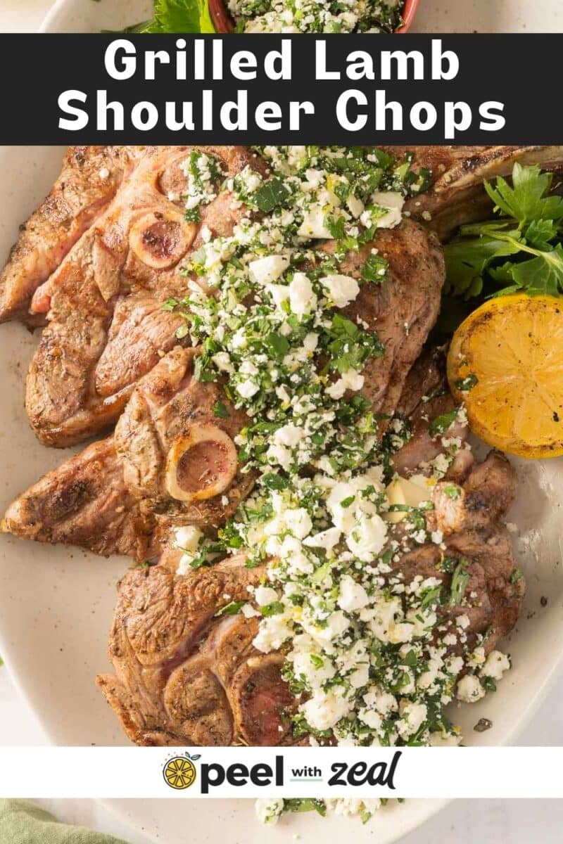 A plate of savory grilled lamb shoulder chops is topped with fresh herbs and creamy feta cheese, all beautifully garnished with a halved lemon.