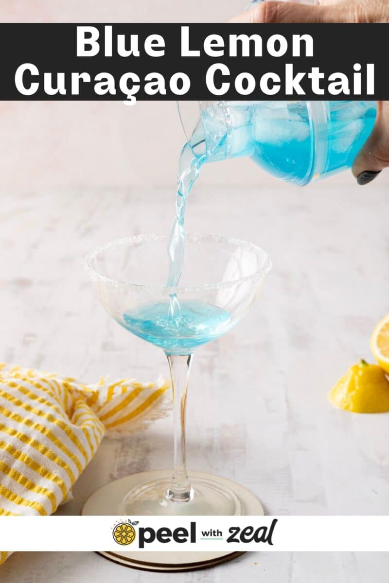Pouring a blue lemon drop martini into a sugar-rimmed glass, with lemon wedges and a yellow-striped cloth on a light surface.