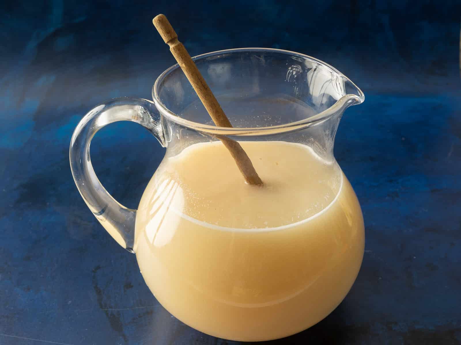 A glass pitcher of horchata with a cinnamon stick stirrer sits elegantly against a blue background, evoking the refreshing allure of lychee lemonade.