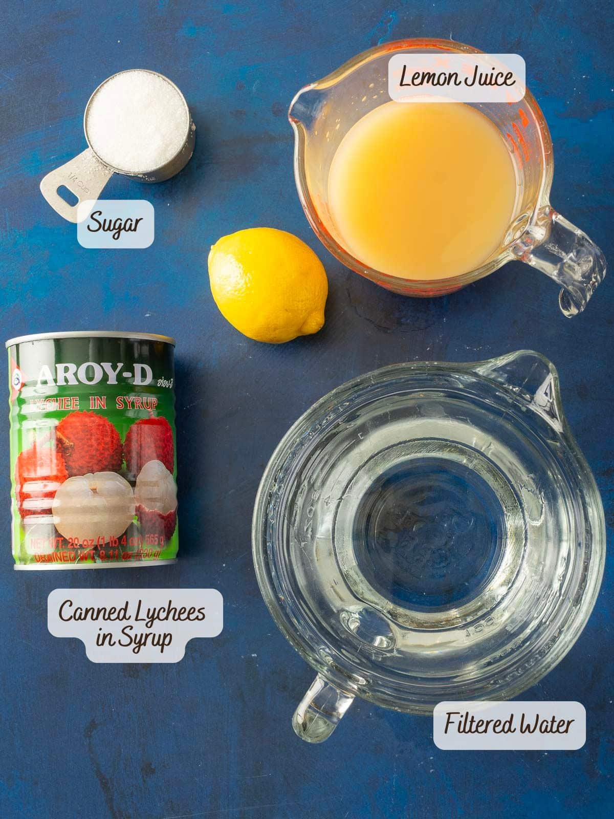 Ingredients for Lychee Lemonade: canned lychees, lemon juice, sugar, lemon, and filtered water on a blue surface.