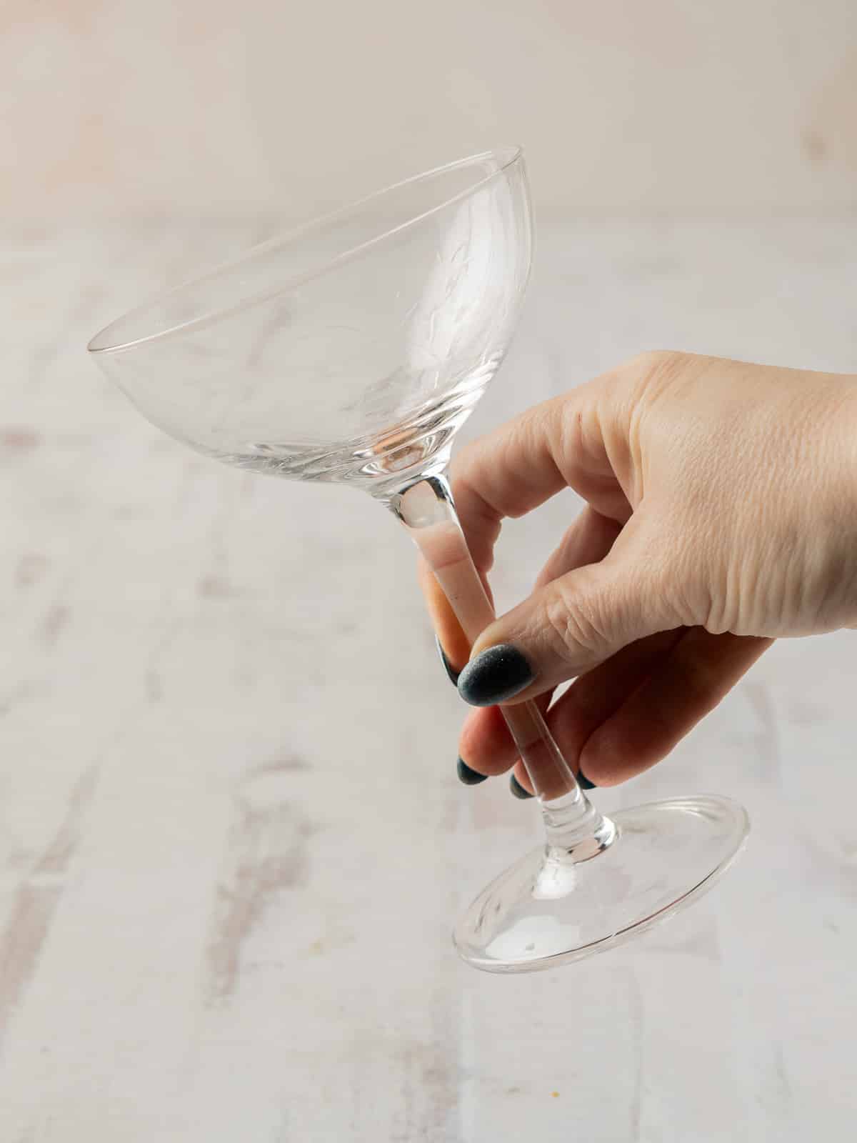 A hand elegantly holds a clear, empty martini glass by its stem on a light surface, inviting thoughts of a refreshing lychee cocktail to come.