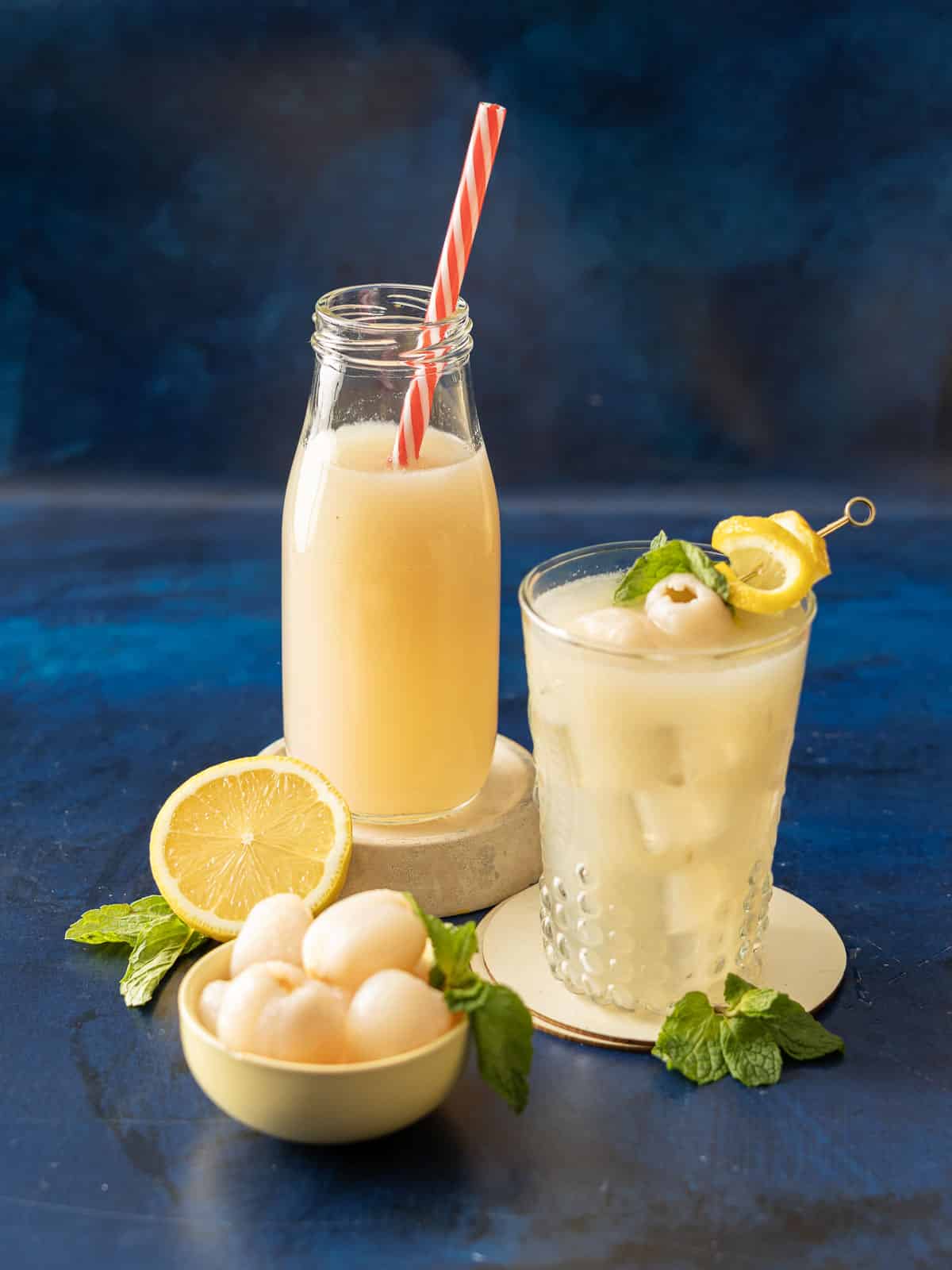 Two refreshing lychee drinks with mint and lemon are set against a blue background. One serves up as a bottle with a straw, the other as a glass brimming with flavor, reminiscent of tangy lemonade.