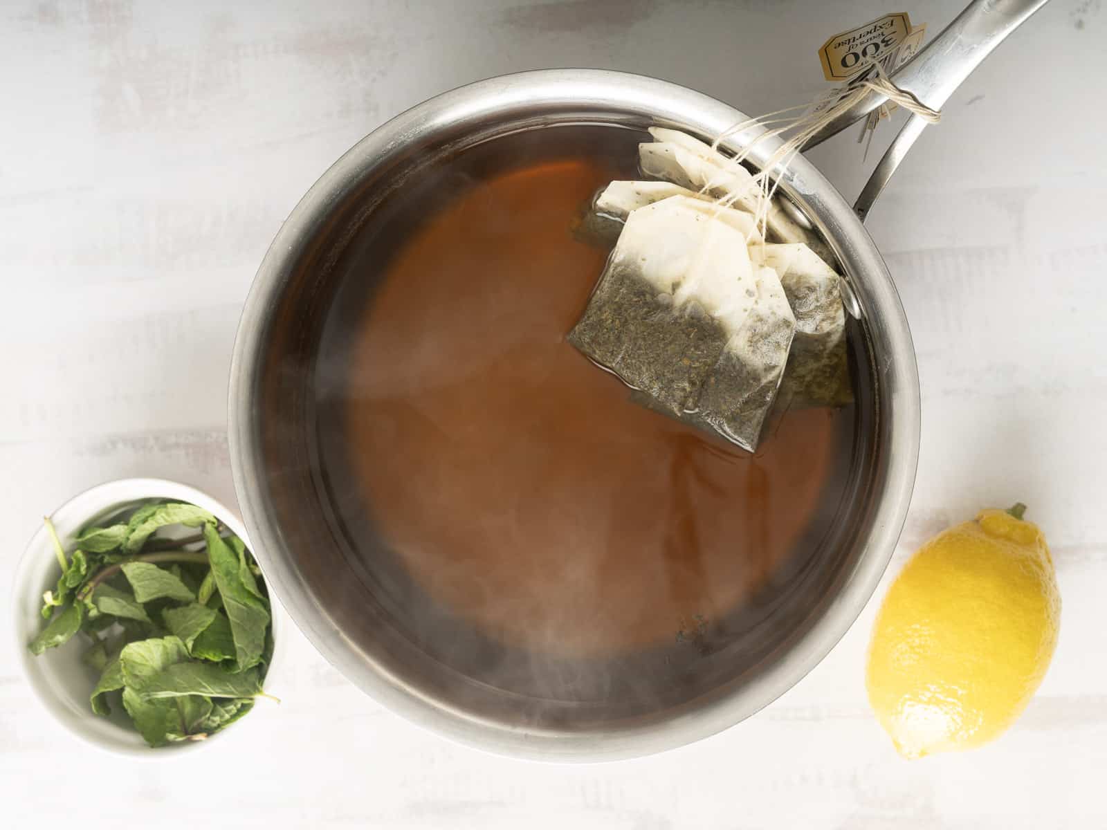 A steaming pot of tea with tea bags, accompanied by a small bowl of mint leaves, a lemon, and hints of lychee iced tea on a pristine white surface.
