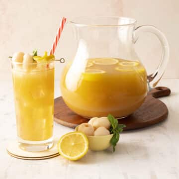 A pitcher of lychee iced tea with lemon slices and a glass with a straw, garnished with mint, lemon, and lychee.