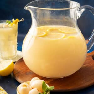 A pitcher of refreshing lemonade with lychee sits surrounded by juicy lychees and a halved lemon, accompanied by a straw-ready glass on a wooden tray.