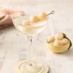 A lychee cocktail elegantly sits in a glass, garnished with skewered lychees, with a bowl of fresh lychees in the background on a light wooden surface.