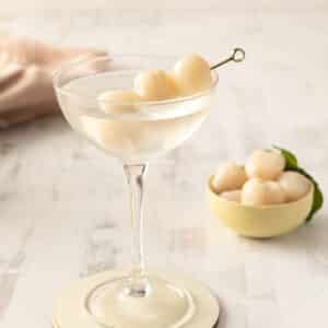 A lychee cocktail elegantly sits in a glass, garnished with skewered lychees, with a bowl of fresh lychees in the background on a light wooden surface.