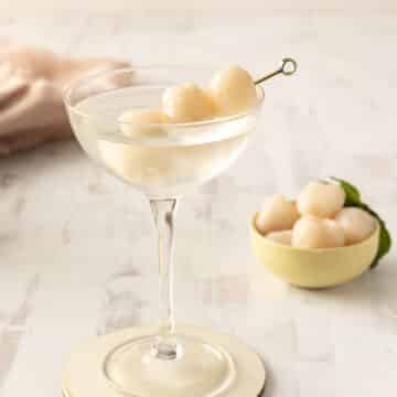 A lychee cocktail elegantly sits in a glass, garnished with skewered lychees, with a bowl of fresh lychees in the background on a light wooden surface.