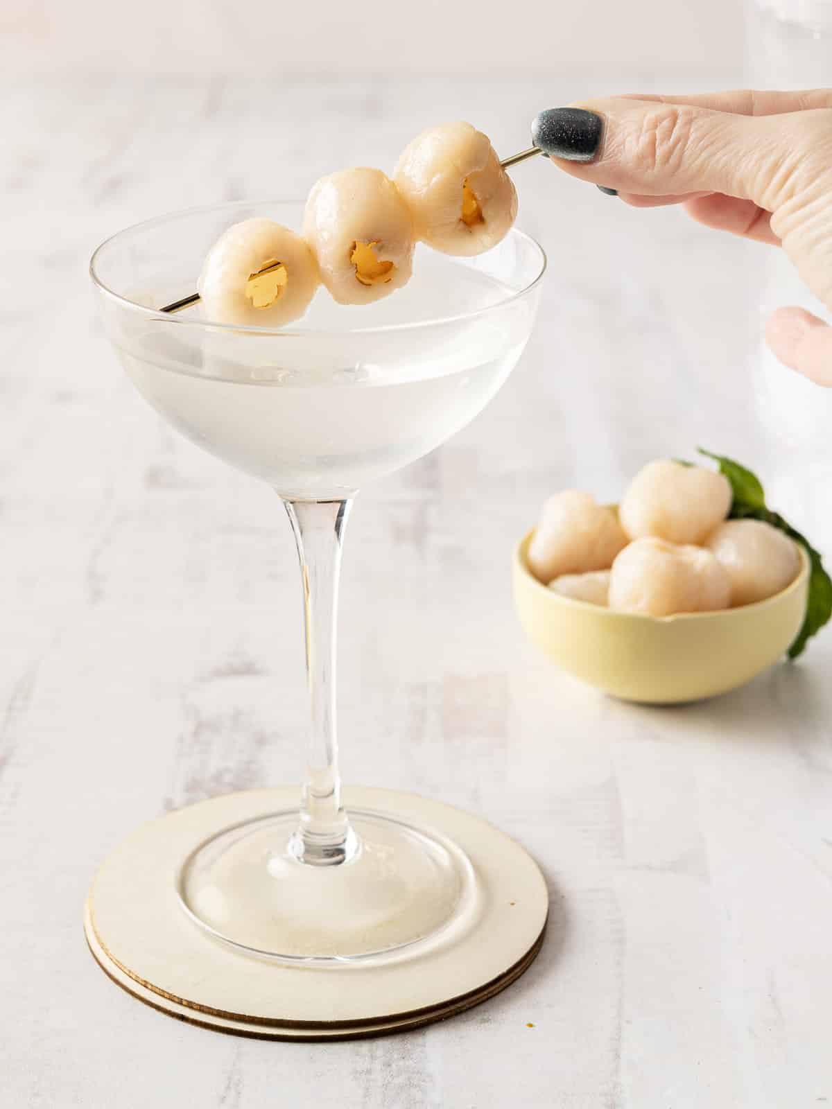 A Lychee Martini sits elegantly in its glass, a hand poised above with a skewer of lychee garnish. In the background, a bowl brimming with fresh lychees adds a touch of intrigue to the delectable cocktail scene.