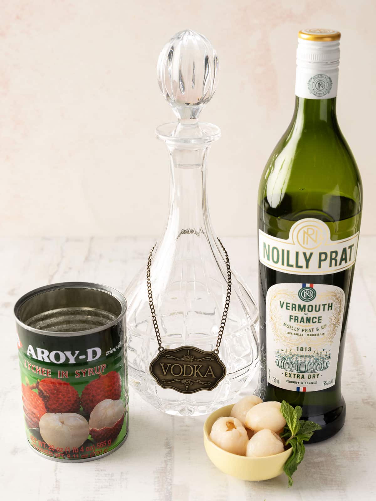 A glass vodka decanter sits elegantly beside a Noilly Prat vermouth bottle, accompanied by a can of lychees and fresh ones scattered nearby. It's the perfect setup for crafting a delightful Lychee Martini.
