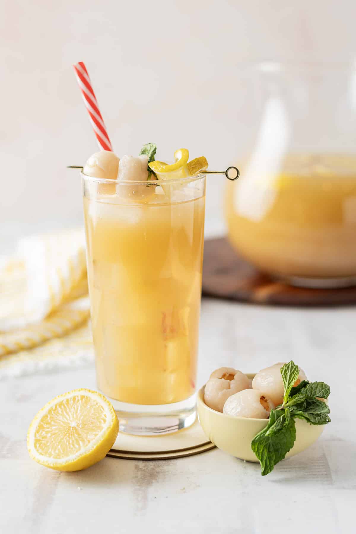 A tall glass of Lychee Iced Tea with ice, elegantly garnished with a juicy lychee, lemon slice, and a fresh mint sprig. A pitcher stands subtly in the background, inviting you to refresh your senses.