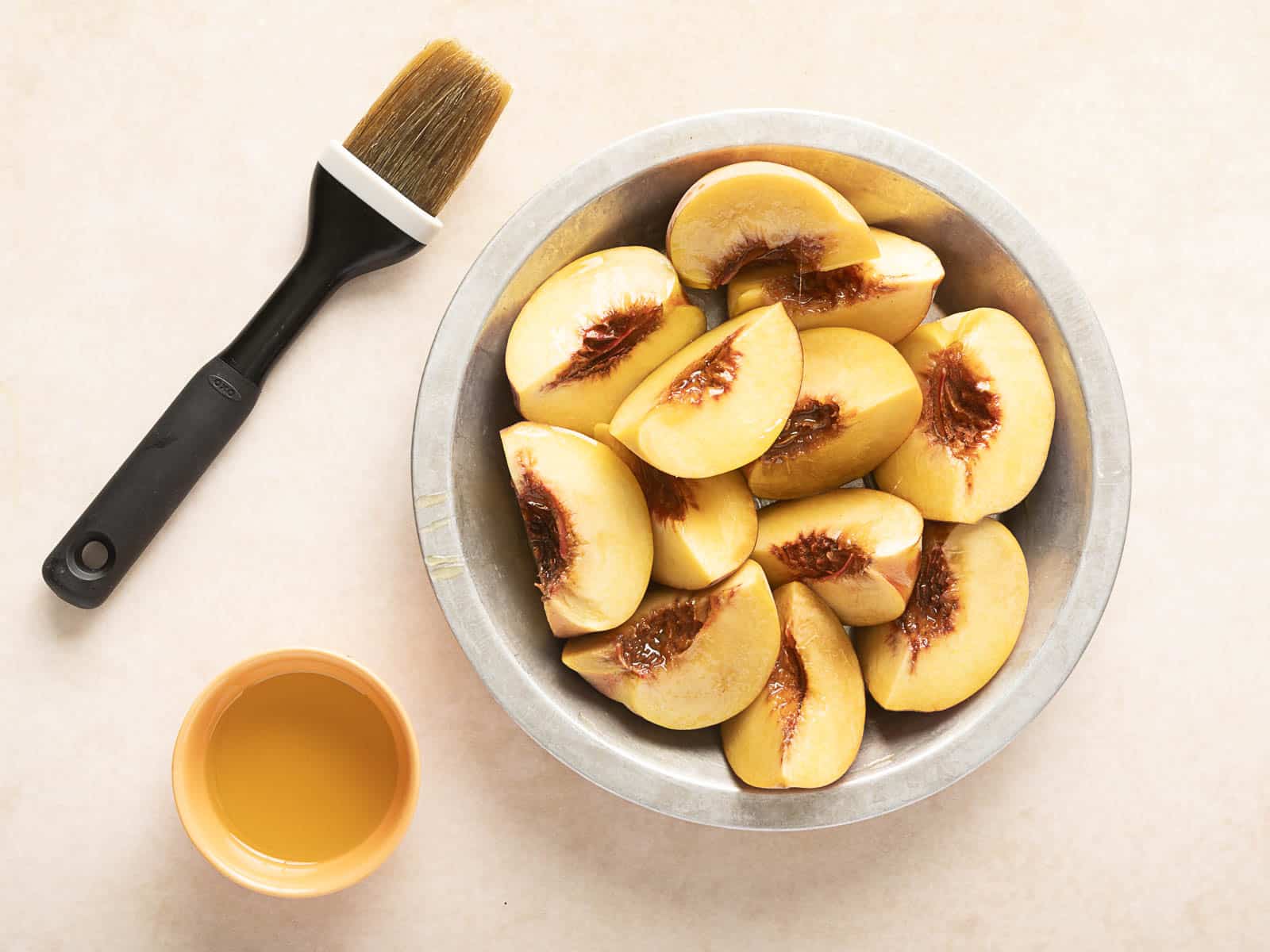 A metal bowl of grilled peach slices sits beside a small container of liquid and a brush on a light surface, ready to complement your salad with burrata.