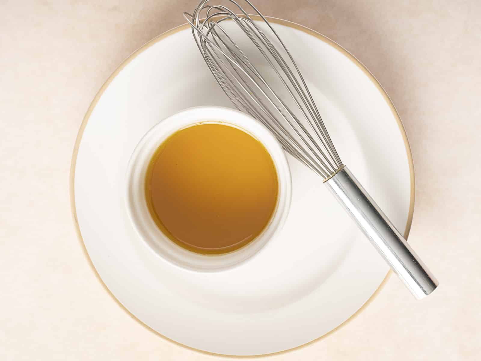 A metal whisk rests on a white plate beside a small bowl filled with golden liquid, viewed from above on a light surface. Nearby, slices of grilled peach promise to elevate the culinary experience.