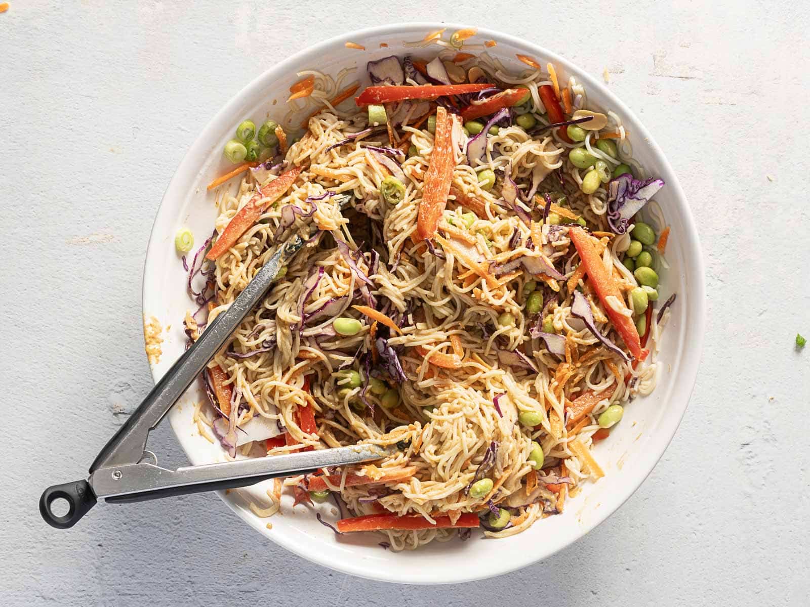 A bowl of cold ramen noodle salad featuring shredded carrots, red cabbage, edamame, and bell peppers, all perfectly mixed and served with tongs.