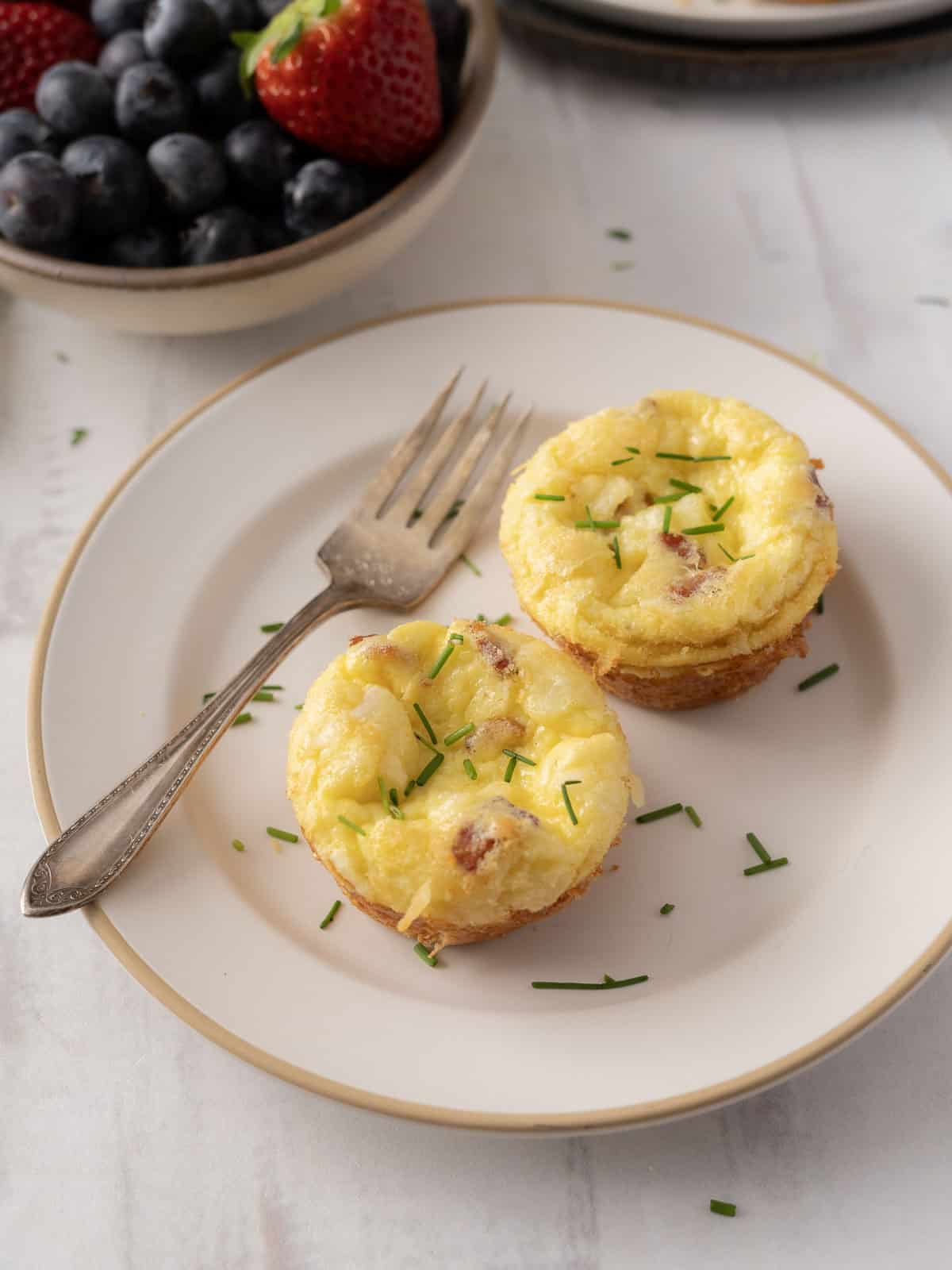 Two quiche muffins with herbs rest on a plate, accompanied by a fork. Berries sit in bowls in the background.