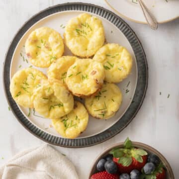 A plate of mini quiche muffins garnished with chives sits next to a bowl of strawberries and blueberries.