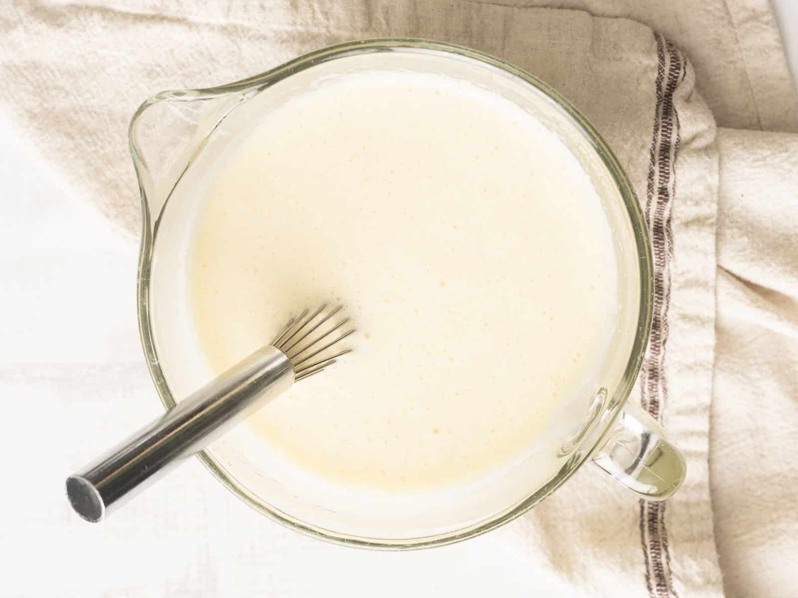 A glass measuring cup with a whisk in creamy quiche batter, placed on a beige cloth.