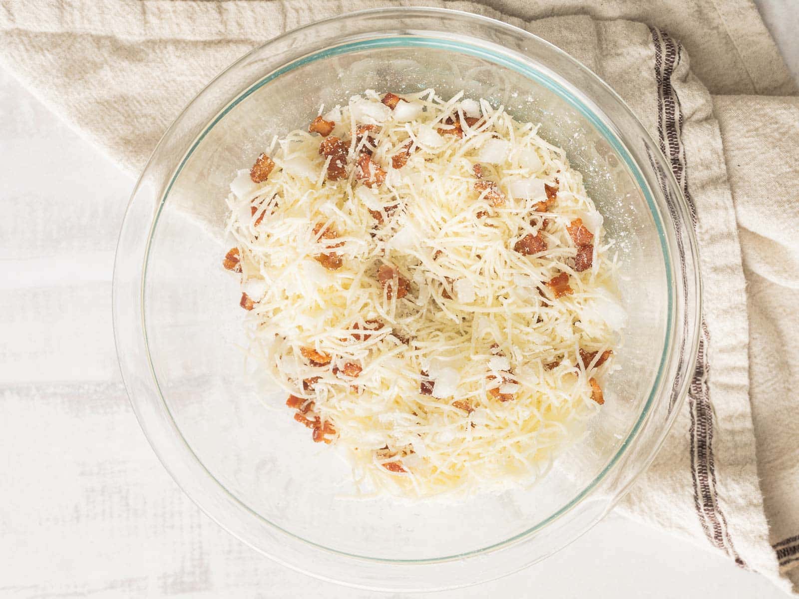 Glass bowl with grated cheese and bacon bits sits delicately on a light cloth napkin, reminiscent of quiche muffin mornings.